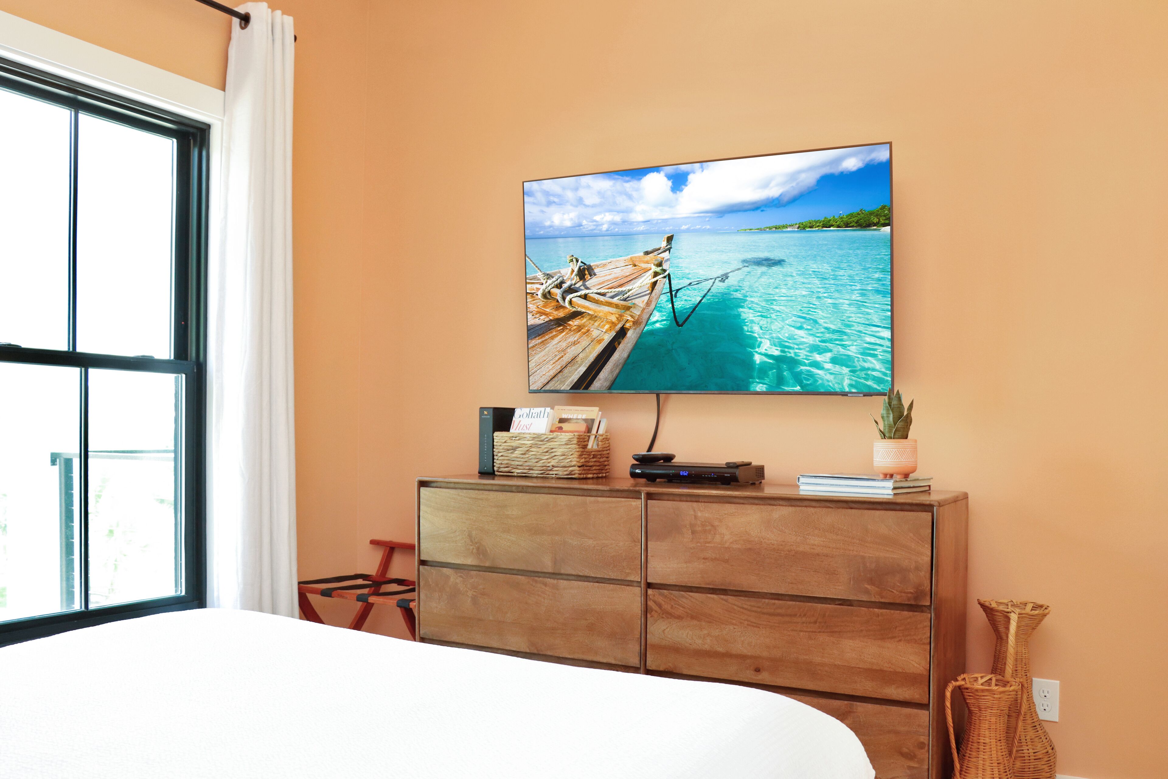 A bedroom with a large window, a wooden dresser, a flat-screen TV mounted on the wall, and a bed.