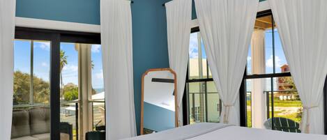 A hotel room with a large bed, a balcony with a view of palm trees, and a window with white curtains.