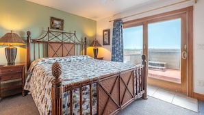 A bedroom with a wooden bed, two bedside tables with lamps, a ceiling fan, a large window with a view of the beach, and a sliding glass door to the balcony.