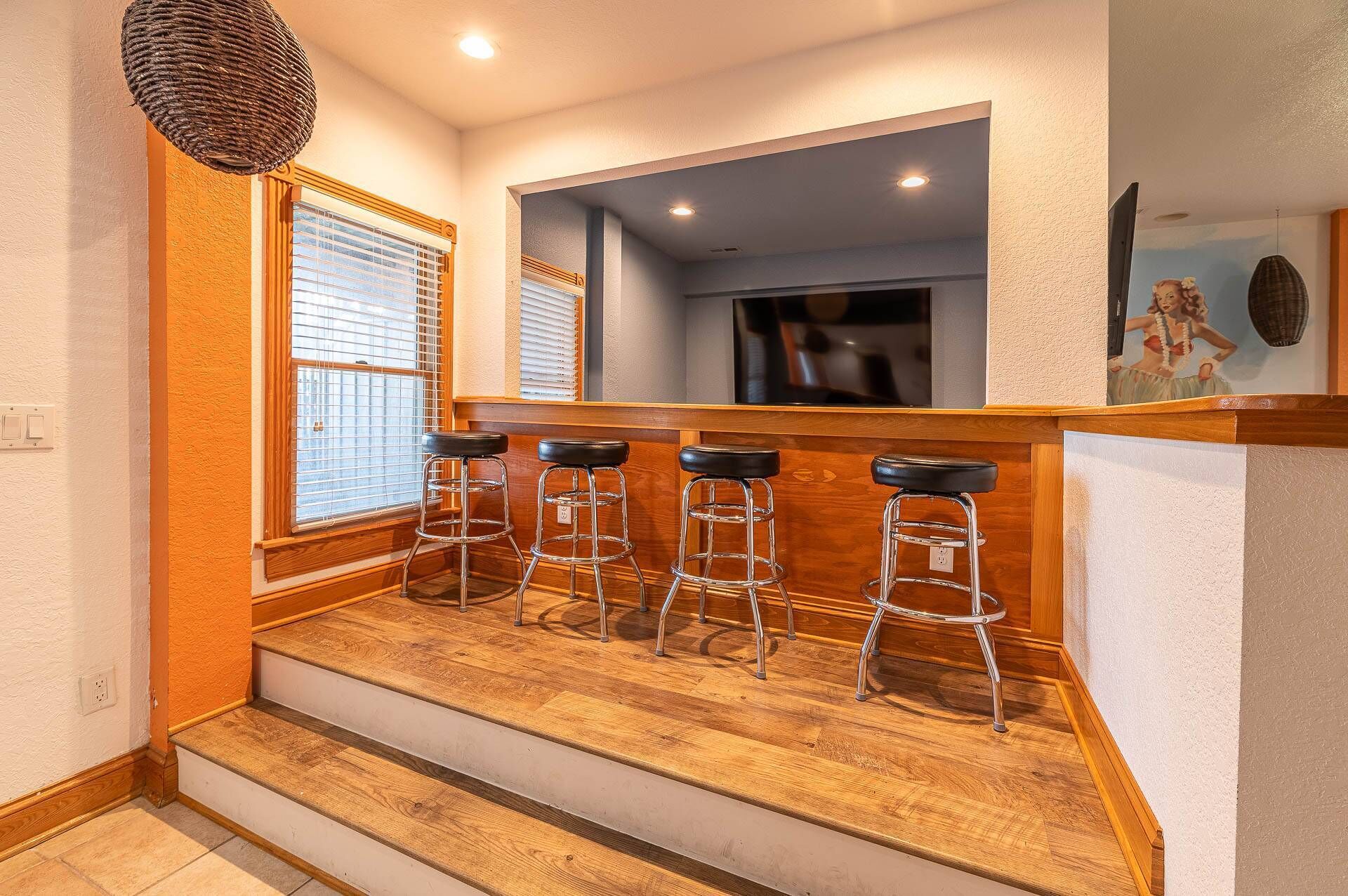 A bar area with four stools, wooden flooring, and a window with blinds.