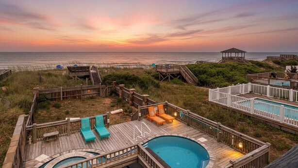 A wooden deck with a pool, lounge chairs, and a gazebo overlooking the beach at sunset.