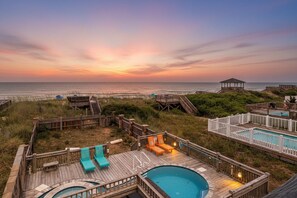 Une terrasse en bois avec une piscine, des chaises longues et un belvédère offrant une vue sur la plage au coucher du soleil.