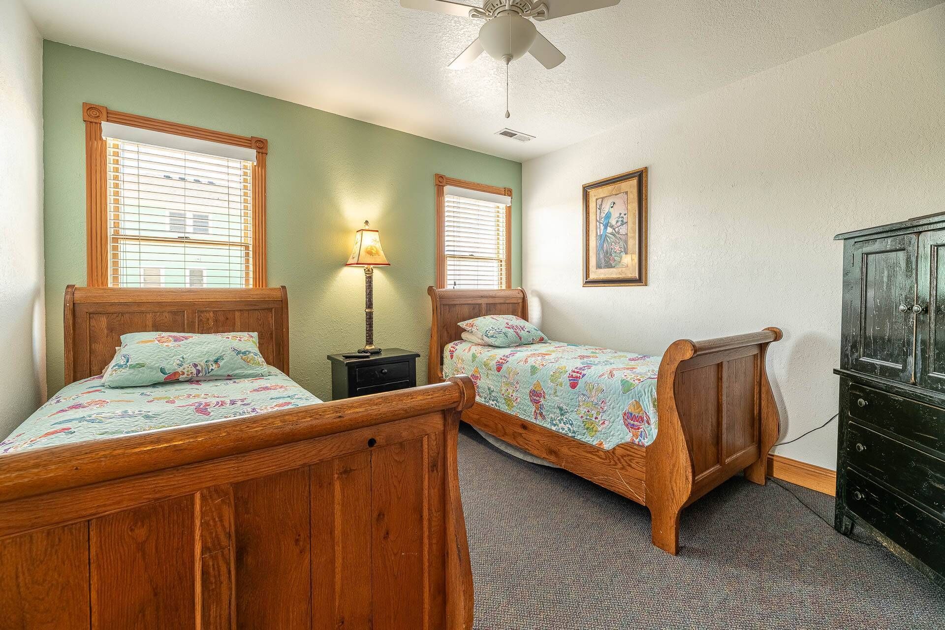 A bedroom with two wooden beds, a nightstand with a lamp, a ceiling fan, and a dresser.