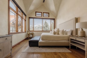 A bedroom with a bed, nightstand, dresser, large windows, and a view of mountains.