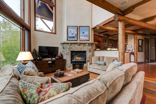 A spacious living room with a fireplace, wooden beams, and a large window overlooking greenery. - Spacious Luxury Home by InvitedHome | Hike-In/Hike-Out, Village Core (Mountain Village)