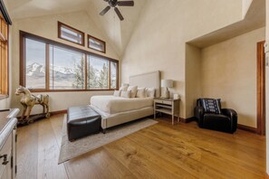 A bedroom with a large window, a bed, a nightstand, a lamp, a chair, and a wooden floor. - Spacious Luxury Home by InvitedHome | Hike-In/Hike-Out, Village Core (Mountain Village)