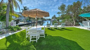 A well-maintained backyard with a dining table and chairs, a thatched umbrella, and a view of a marina with boats.