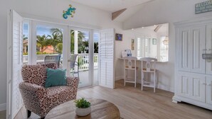 A room with a patterned sofa, a small round table with a plant, a dining area with white chairs, and a view of palm trees outside.