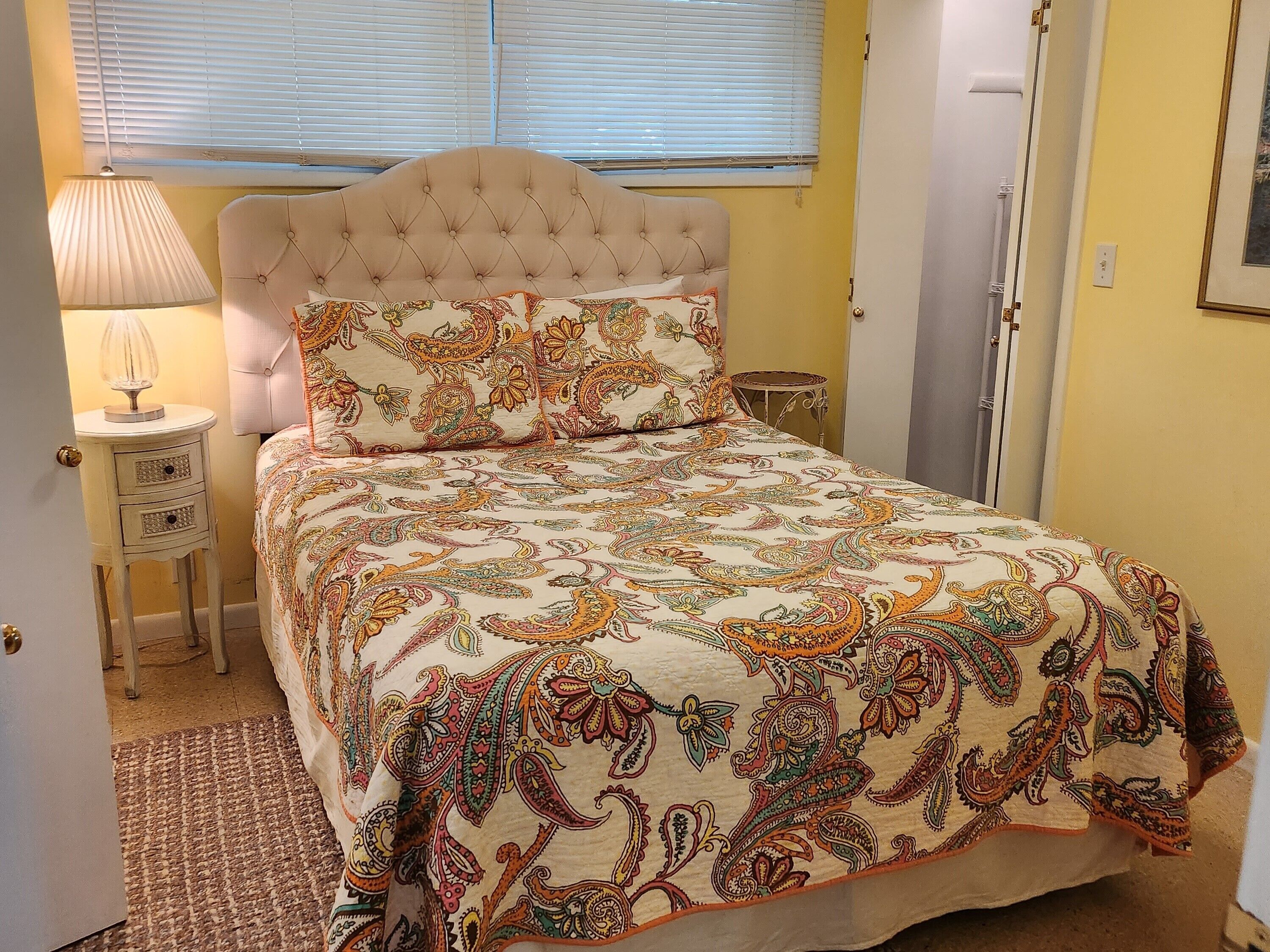 A bedroom with a tufted headboard, a patterned bedspread, a bedside lamp, and a small table.