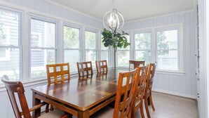 Une salle à manger avec une grande table en bois et six chaises, de grandes fenêtres et un luminaire suspendu.