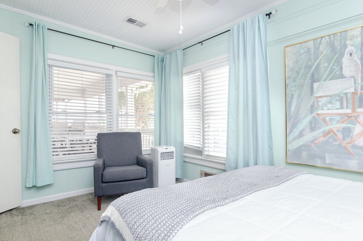 A bedroom with a bed, a gray armchair, a window with blinds, and a painting on the wall.