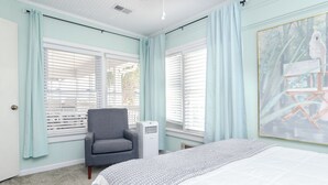 Une chambre à coucher avec un lit, un fauteuil gris, une fenêtre avec des stores et un tableau accroché au mur.