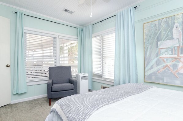 A bedroom with a bed, a gray armchair, a window with blinds, and a painting on the wall.