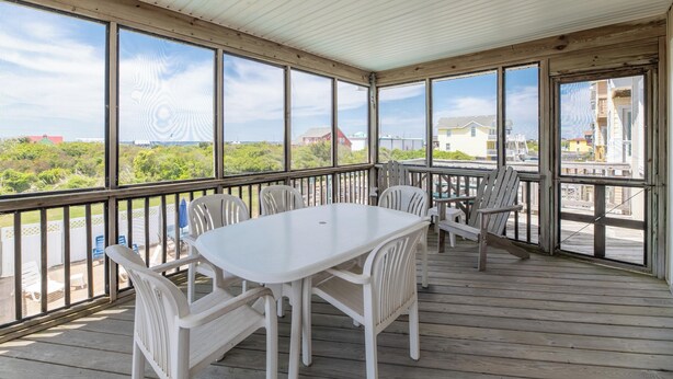A covered outdoor deck with a white table and chairs, large windows, and a view of greenery and buildings. - Fun 'N Sun - Nags Head MP 16, 250 Ft to Beach Access, Pool, Pets OK, YR Flex Stays (Nags Head)