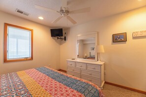 Une chambre à coucher comprenant un lit, une commode, un ventilateur de plafond, une télévision, une fenêtre avec des stores et des éléments de décoration murale.
