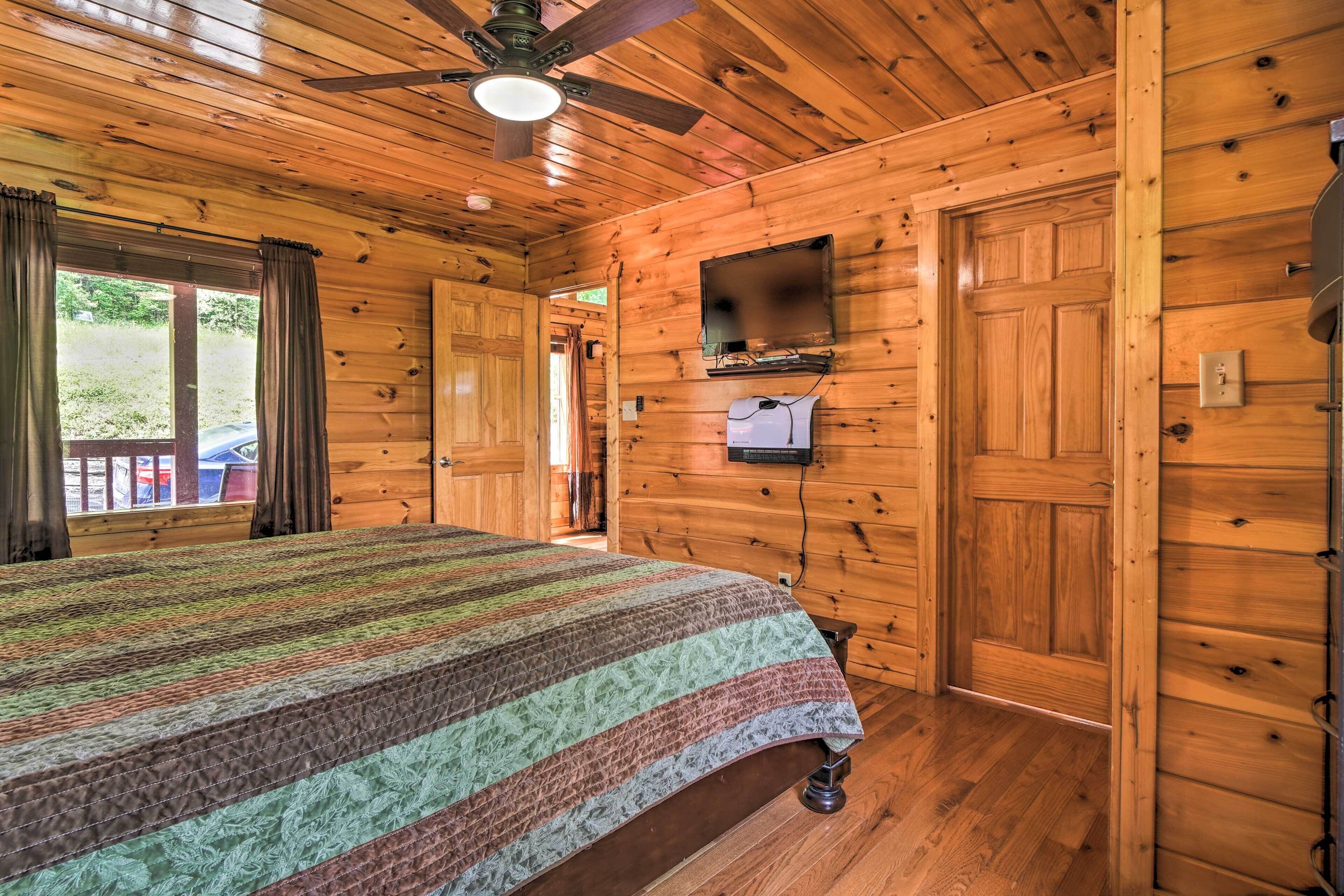 A wooden cabin room with a bed, a ceiling fan, a TV mounted on the wall, and a door leading outside.