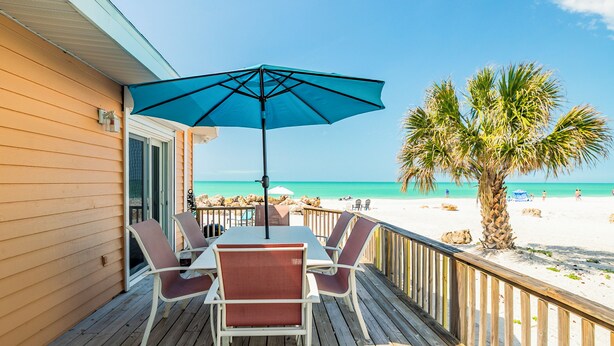 Eine Holzterrasse mit Tisch und Stühlen, einem blauen Sonnenschirm und Blick auf Strand und Palmen.