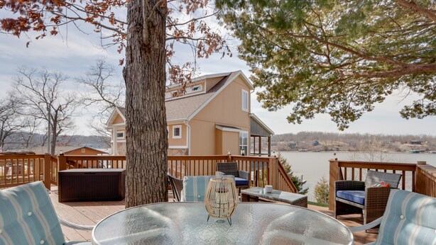 Una terraza con mobiliario exterior, una mesa de vidrio y vistas a una casa y un lago.