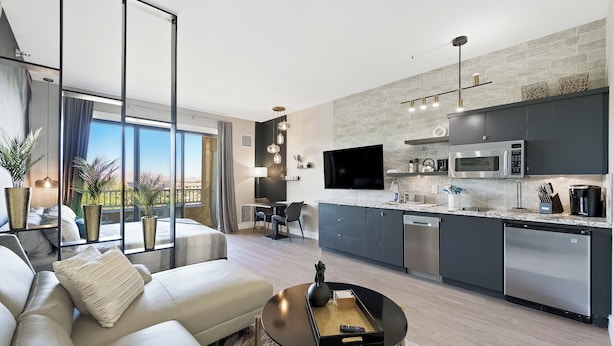 A modern living room with a sofa, a coffee table, and a TV. The kitchen has a microwave, a dishwasher, and a countertop with various items.