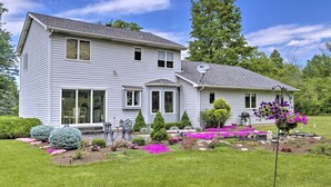 Une maison à deux étages avec un porche à l’avant, une antenne parabolique et un jardin bien entretenu où poussent différentes plantes et fleurs.