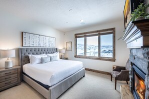 A bedroom with a large bed, a stone fireplace, a window with a snowy view, and a framed artwork on the wall.