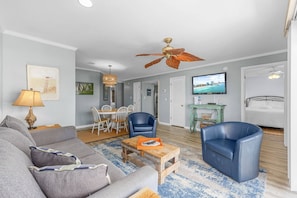 Un salon comprenant un canapé, deux fauteuils bleus, une table basse en bois et un ventilateur de plafond. On y trouve également un coin repas avec une table et des chaises, ainsi qu’un téléviseur fixé au mur.