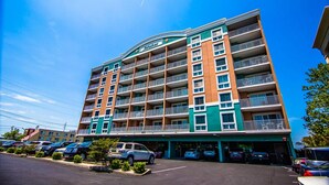 A multi-story hotel with a green and beige facade, multiple balconies, and a parking lot in front. - Bayfront unit. Walk to the Convention Center, dining, shops and the beach. (Ocean City)