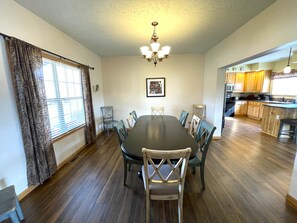 A dining room with a large table and chairs, a chandelier, a window with curtains, and a kitchen area with wooden cabinets and appliances.