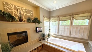 A bathroom with a bathtub, a TV mounted on the wall, a fireplace, and a window with shutters.