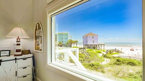 A room with a large window overlooking a beach, a lighthouse, and a wooden structure on stilts.