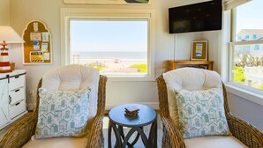 A room with wicker chairs, a small table, a TV, and a window with a beach view.
