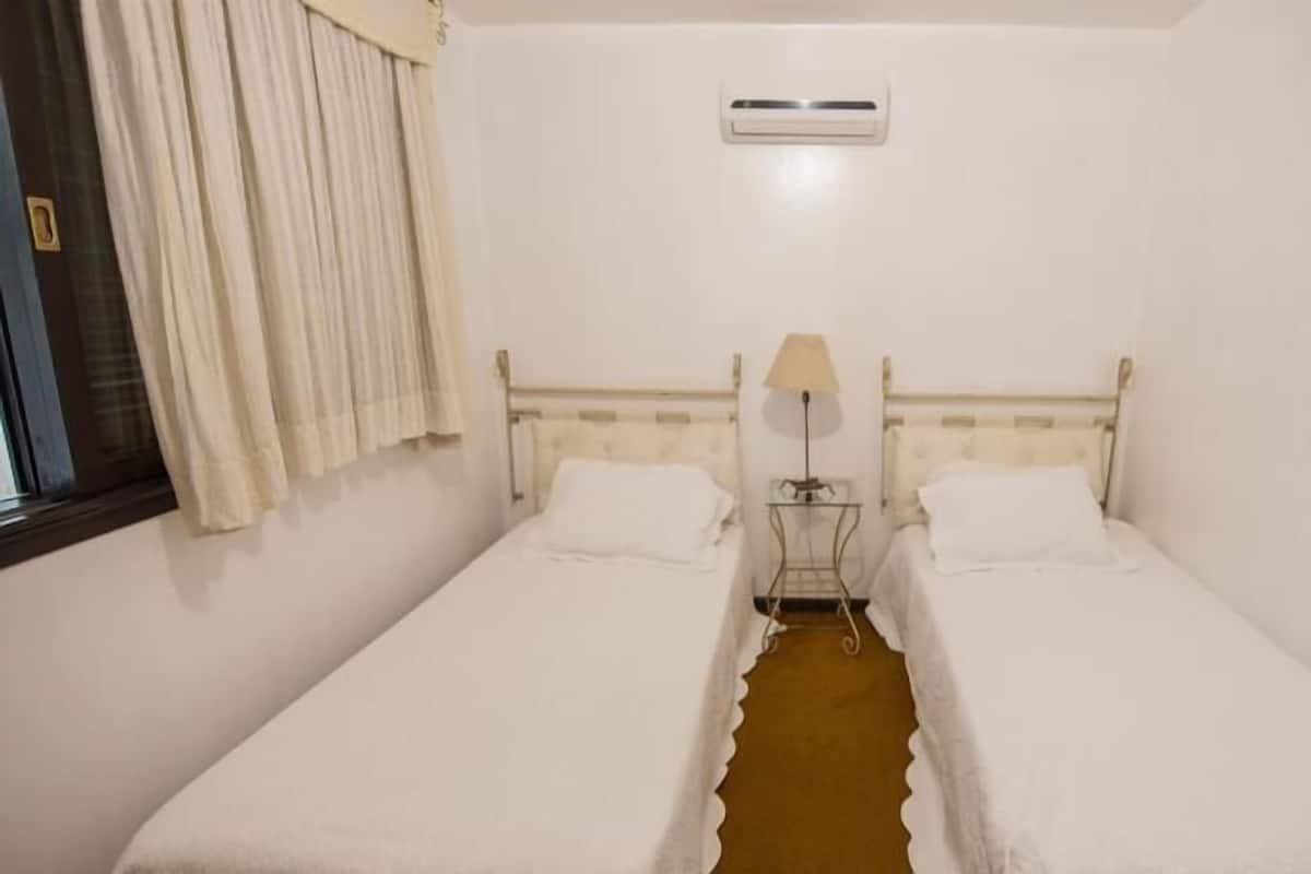 Une petite chambre propre, avec deux lits simples, une table de chevet avec une lampe et une fenêtre avec des rideaux.