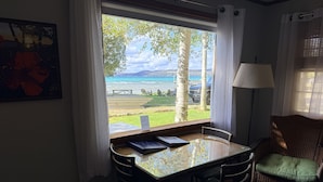 A room with a view of a lake, a table with chairs, a wicker chair, a lamp, and a framed picture on the wall. - Cozy cottage Big Glen Lake, Tennis Court, Dock, Kayaks, Rowboat, Sunset view (Maple City)