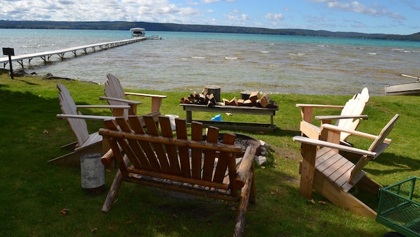 A lakeside deck with Adirondack chairs, a fire pit, and a dock extending into the water. - Cozy cottage Big Glen Lake, Tennis Court, Dock, Kayaks, Rowboat, Sunset view (Maple City)