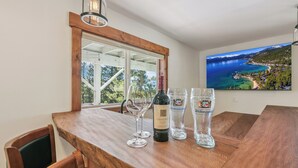 A wooden bar with a wine bottle and glasses, a large window with a view of a coastal landscape, and a mounted painting of the same view. - Heavenly Mountain View: Pool Table, Hot Tub, BBQ (South Lake Tahoe)