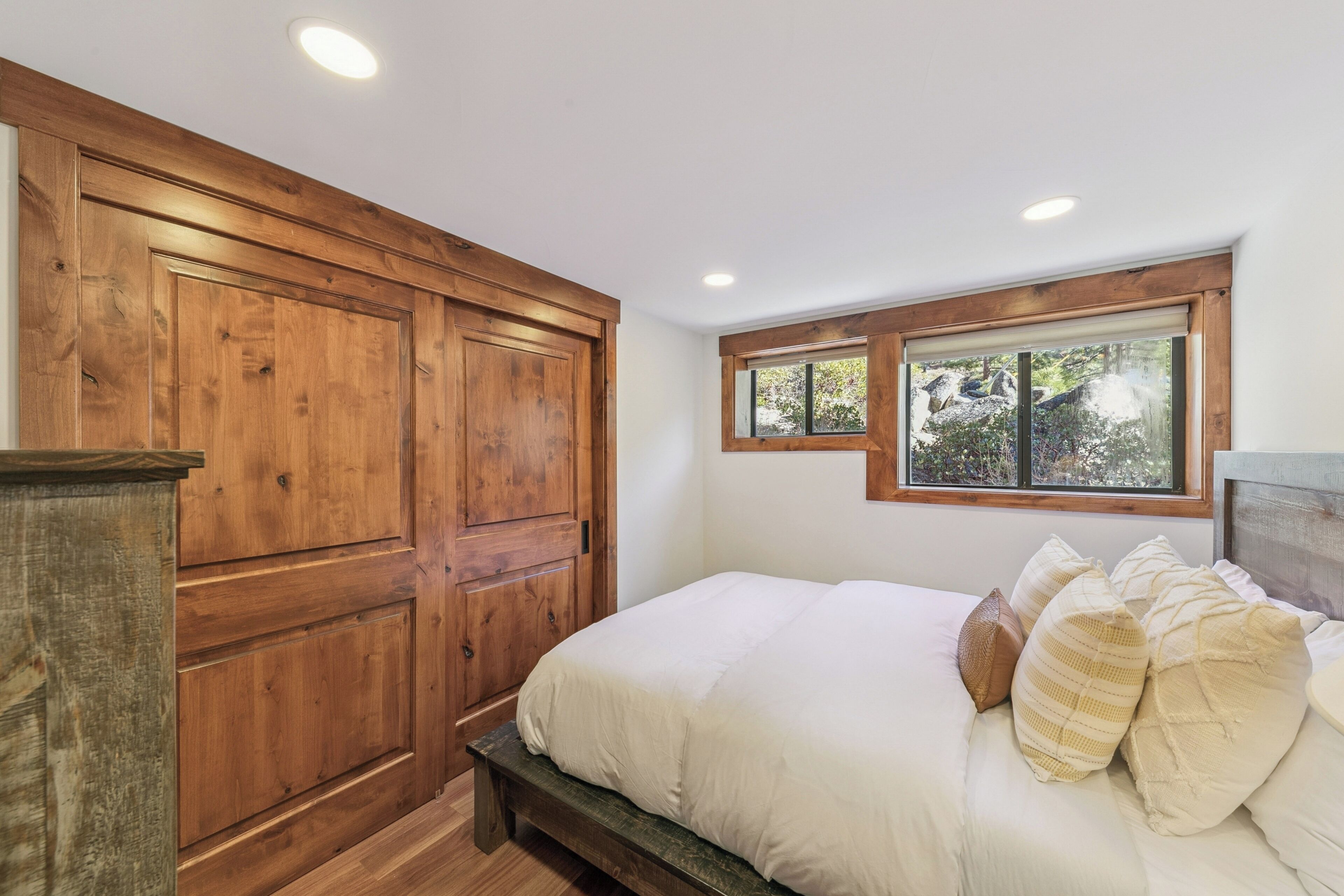 A bedroom with a wooden wardrobe, a bed with white bedding, and a window with a view of trees.
