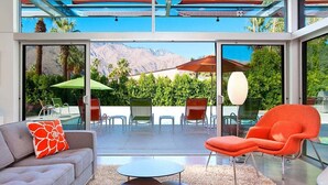 A modern living room with a sofa, armchair, and round tables, overlooking a pool and mountains.