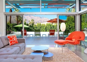 A modern living room with a sofa, armchair, and round tables, overlooking a pool and mountains.