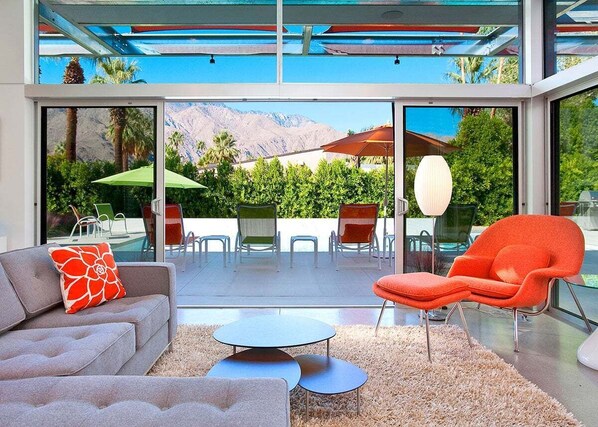 Sala moderna com sofá, poltrona e mesas redondas, com vista para uma piscina e montanhas.