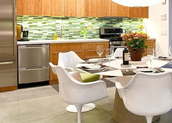 Una cocina moderna con armarios de madera, un refrigerador de acero inoxidable y un comedor con sillas blancas y una mesa dispuesta para dos personas.