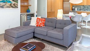 A modern living room with a grey sectional sofa, a coffee table, and a dining area with white chairs.