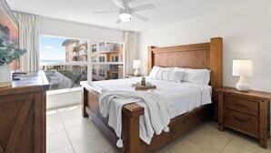 Un dormitorio con una cama de madera, dos mesitas de noche con lámparas, un gran ventanal con vistas a edificios y un ventilador de techo.