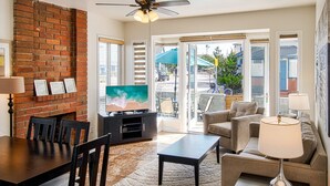 A living room with a brick wall, a ceiling fan, a TV, a dining table with chairs, a sofa, and a coffee table. - Awesome lower unit in a duplex just one house away from the sand and boardwalk! (Newport Beach)
