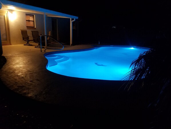 A swimming pool with a blue light, a covered patio area, and a few chairs.