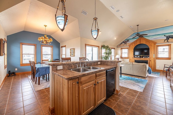 A spacious kitchen with a central island, wooden cabinets, and a built-in dishwasher. There is a dining area with a blue table and chairs, and a living room with a sofa and a fireplace.
