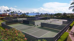 Ein Tennisplatzkomplex mit mehreren Plätzen, umgeben von Grünflächen und Palmen, mit Blick auf den Ozean im Hintergrund.