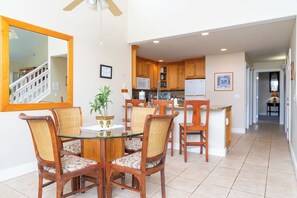 A dining area with a glass table and wicker chairs, a kitchen with wooden cabinets, and a hallway leading to a bedroom. - 3 bed/3 bath Poipu Condo - Central A/C, Walking path to Beach (Koloa)