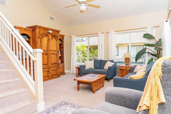 A living room with a staircase, a wooden cabinet, a sofa, a coffee table, and a window with curtains. - 3 bed/3 bath Poipu Condo - Central A/C, Walking path to Beach (Koloa)