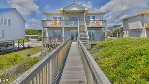 Una casa affacciata sulla spiaggia con più piani, dotata di una veranda in legno che conduce all'ingresso, un balcone con mobili da esterno e una vista libera sull'oceano.