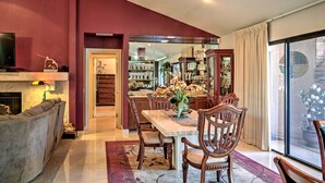 Un coin repas avec une table dressée pour quatre personnes, entouré de chaises en bois, d’une cheminée et d’une porte vitrée donnant sur un espace cuisine avec des étagères remplies de vaisselle et une plante en pot.